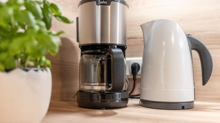 Een moderne koffiemachine staat naast een waterkoker op een houten oppervlak. Op de achtergrond is een pot met verse kruiden te zien.