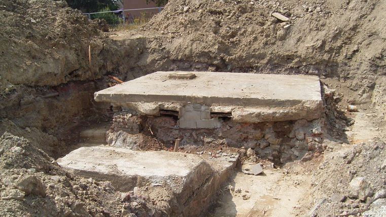 Ein ausgehobenes Grundstück mit einem Betonfundament. Rund um das Fundament liegen erhobene Erdschichten.