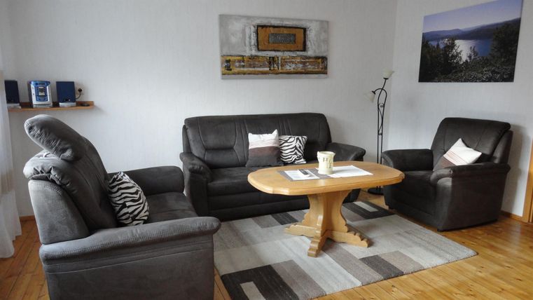 Een gezellige woonkamer met een grijze bankenset en een ronde houten tafel. Aan de muur hangt een schilderij en een klein rek is versierd met luidsprekers.