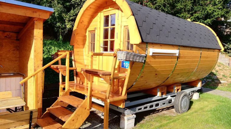 Ein gemütliches Holzfass mit einer überdachten Veranda und großen Fenstern. Es steht auf einem Anhänger und ist von grünem Gras umgeben.