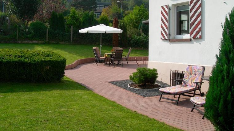 A beautiful garden with a terrace and garden furniture. In the background, there is a sun umbrella and a well-maintained lawn.
