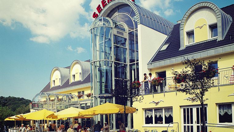 Een modern restaurant met een grote glazen façade en gele parasols. Op de voorgrond zitten gasten buiten en eten.