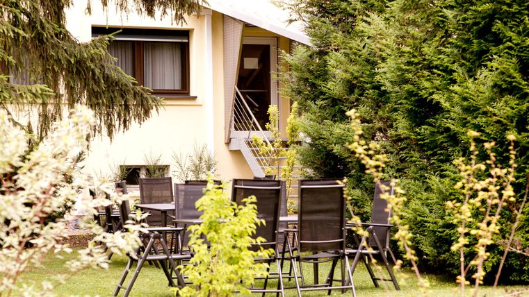 A well-maintained garden with a set of chairs and tables. In the background, a house and tall plants can be seen.