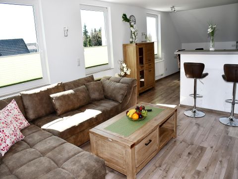Ein helles Wohnzimmer mit einem beigen Sofa und einem Holztisch. Im Hintergrund befindet sich eine moderne Küchenzeile.