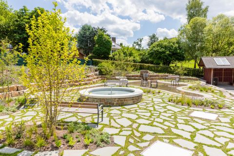 Ein schöner Garten mit einem runden Brunnen und Sitzbänken. Die Umgebung ist grün mit Bäumen und Sträuchern, und der Himmel ist teilweise bewölkt.