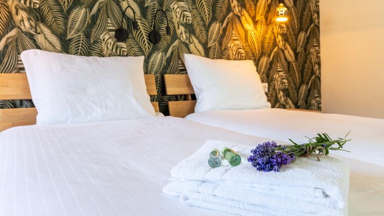 A cozy bedroom with two beds and an elegant plant wallpaper. Freshly folded towels and a small bouquet of lavender are decoratively placed on the bed.