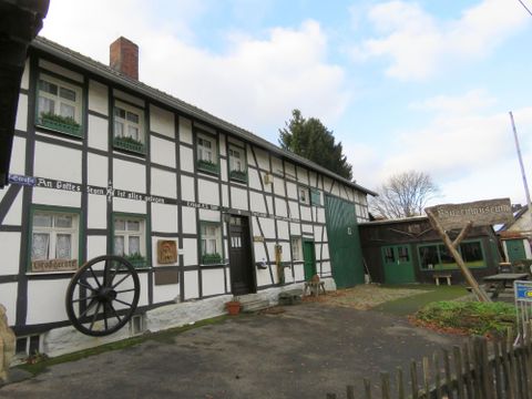 A traditional half-timbered house with white walls and dark beams. In the foreground, there is a large wagon wheel, and the garden is well-kept.
