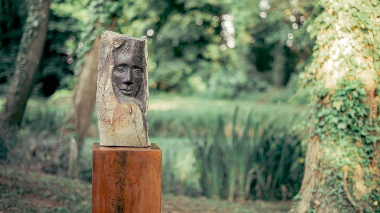 Een houten sculptuur met een gezicht staat op een sokkel in een groene park. Op de achtergrond zijn bomen en een rustige landschap te zien.