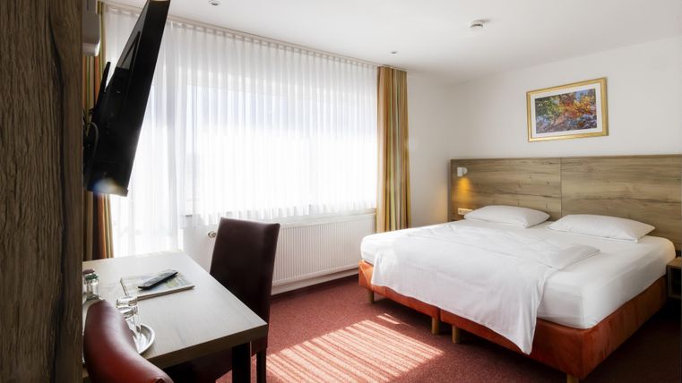 Ein helles Hotelzimmer mit einem großen Doppelbett und einem Schreibtisch. Das Zimmer hat große Fenster, die viel Tageslicht hereinlassen.