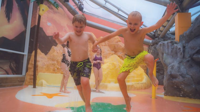 Twee blije kinderen springen in een waterpark. Op de achtergrond zijn kleurrijke speeltoestellen en een ontspannen vakantiesfeer te zien.