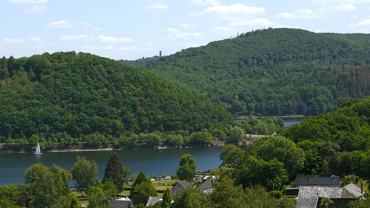 Eine malerische Landschaft mit grünen Hügeln und einem klaren Fluss. Im Vordergrund sieht man kleine Häuser und einen Segelboot auf dem Wasser.