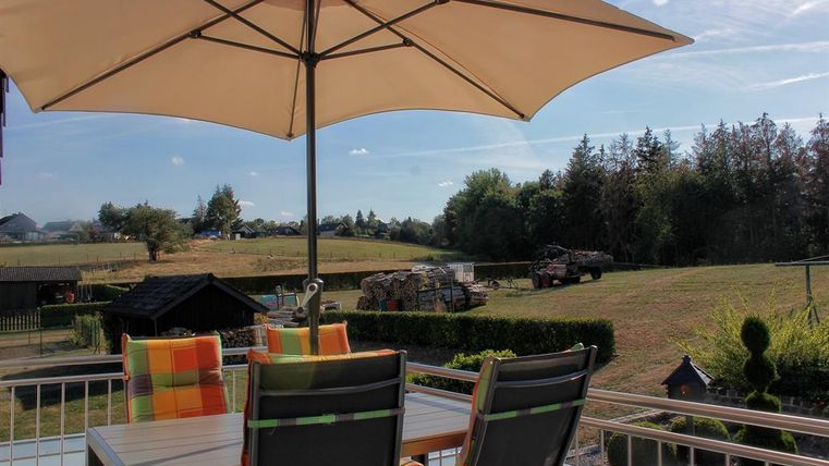 Een gezellig balkon met een parasol en een tafel. Op de achtergrond zie je een groene weide en bomen.