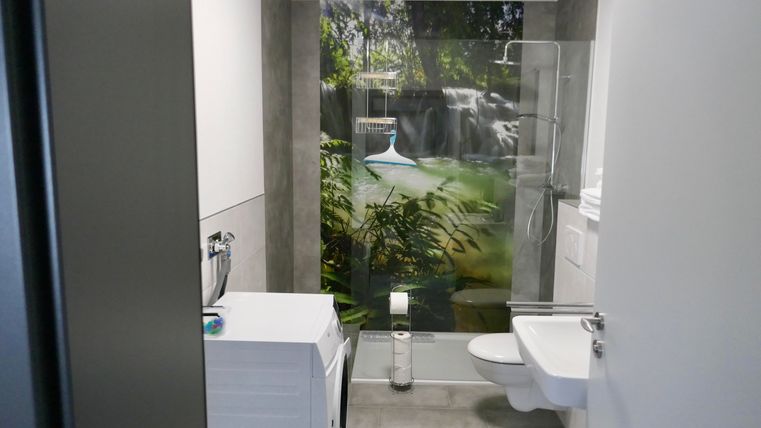 A modern bathroom with a washing machine and a bright, inviting environment. A wallpaper featuring a beautiful waterfall creates a relaxing atmosphere.