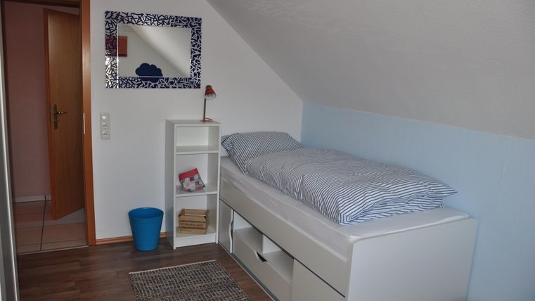 A cozy bedroom with a single bed and a bright wall. The room includes a shelf and a blue trash can.