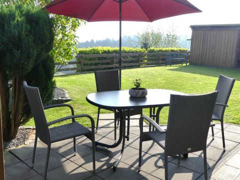 An inviting outdoor area with a round table and four chairs under a red sun umbrella. The garden is green and well-maintained, perfect for relaxed hours outdoors.