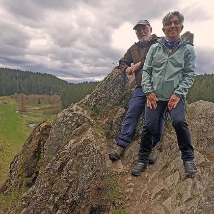 Ein Paar steht auf einem Felsen in der Natur. Im Hintergrund sind grüne Wälder und ein bewölkter Himmel zu sehen.