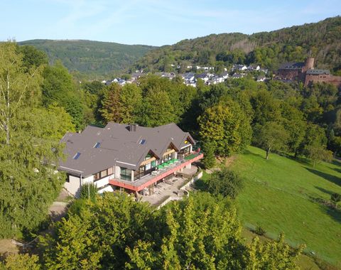 A cozy hotel surrounded by green trees and gentle hills. In the background, houses and a castle are visible.