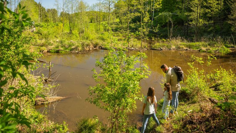 A family stands by the shore of a tranquil body of water, surrounded by lush greenery. The sun is shining and nature is alive.