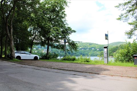 A quiet street with trees and a white car. In the background, a lake and rolling hills can be seen.
