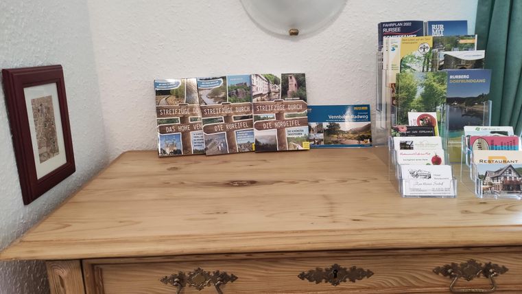 A wood-colored table with a small selection of postcards and informational materials. A framed picture hangs on the wall in the background.