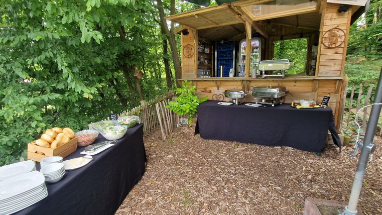A cozy outdoor dining area with a wooden stand and a buffet. Fresh rolls, salads, and dishes are arranged on a table, surrounded by trees.