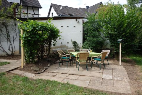 Een gezellige zithoek in de tuin met een ronde tafel en stoelen. Omringd door planten en een witte muur.