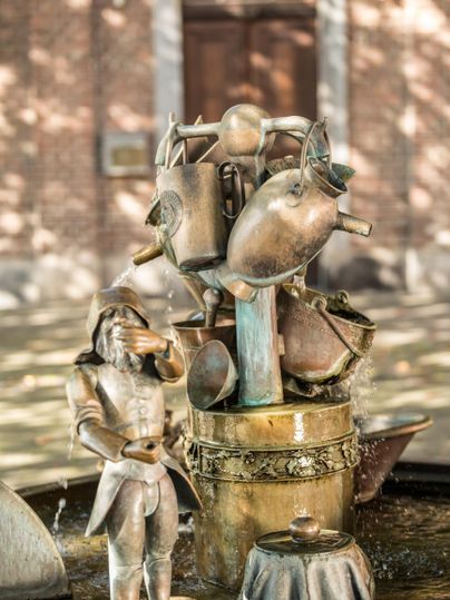 An artistic bronze sculpture featuring a soldier and several jugs. The sculpture stands in a fountain and reflects sunlight.