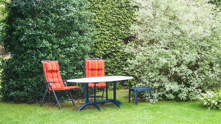 A beautiful garden with a table and two chairs in vibrant colors. Surrounded by green shrubs and lawn.
