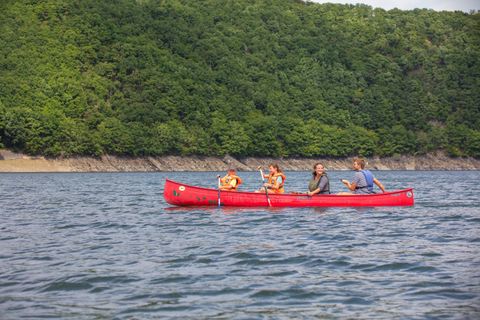 Eine Gruppe von vier Personen paddelt in einem roten Kanu auf ruhigem Wasser. Im Hintergrund ist eine grüne, bewaldete Küste zu sehen.