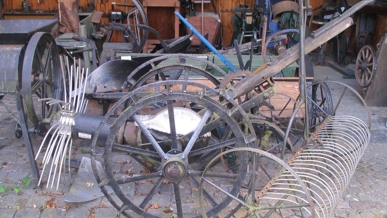 A collection of old agricultural tools and machines. The objects are made of metal and wood and are located in a barn.