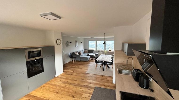 A modern kitchen with wooden flooring and elegant appliances. In the background, there is a cozy living area with a dining table and a beautiful view.