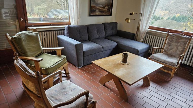 Ein gemütliches Wohnzimmer mit einem grauen Sofa und einem Holztisch. Rundherum stehen mehrere Stühle und große Fenster bieten Blick auf die Natur.