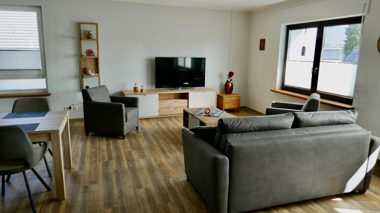 A modern living room with a grey couch, two armchairs, and a television. The room has large windows and wooden flooring.