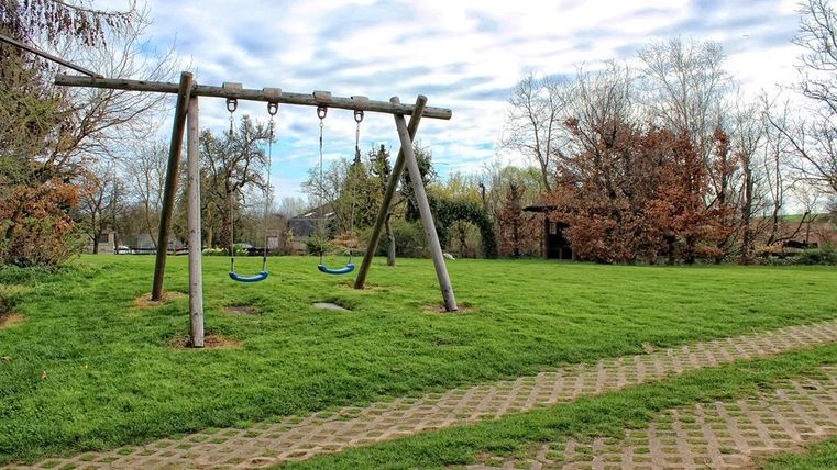 Een speelplaats met twee schommels staat op een groene weide. Op de achtergrond zijn bomen en een pavilion te zien.