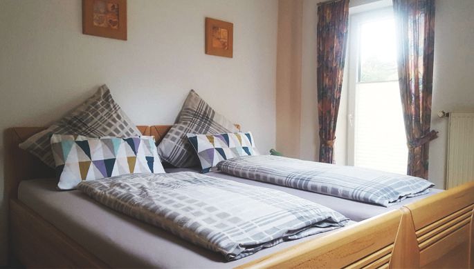 A cozy bedroom with two single beds and colorful cushions. The window lets in plenty of daylight and the curtains are colorful.
