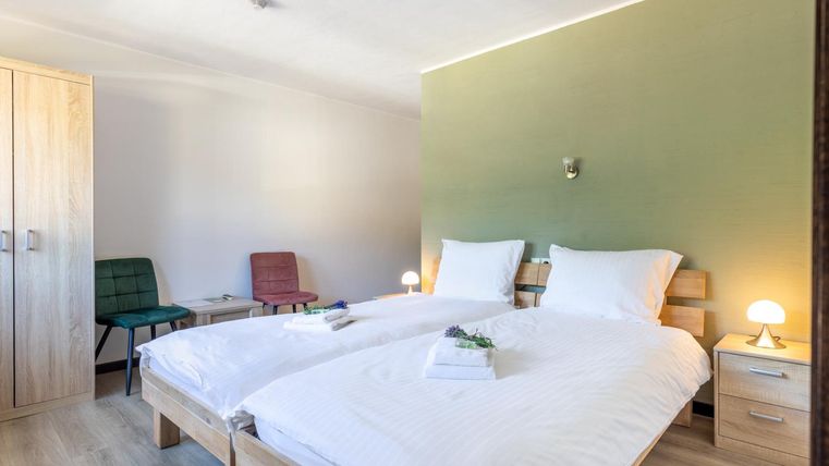 A modern bedroom with two single beds and wooden ceilings. The room features a calming green wall and cozy chairs.