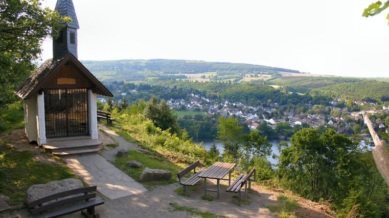 Een schilderachtig uitzicht vanaf een heuvel met een kleine kapel. Op de achtergrond strekt zich een dorp langs een rivier uit, omgeven door groene heuvels.