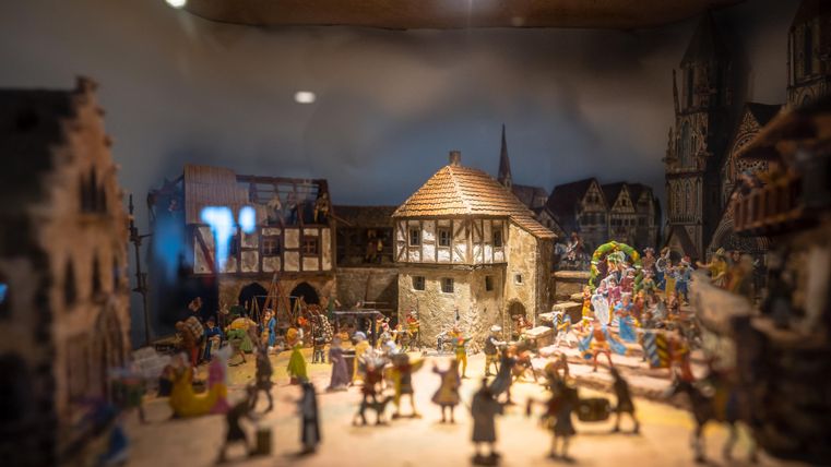 Een gedetailleerde miniatuurscène met een dorp en figuren die een feestelijke sfeer creëren. De afbeelding toont historische gebouwen en blije mensen te midden van activiteiten.