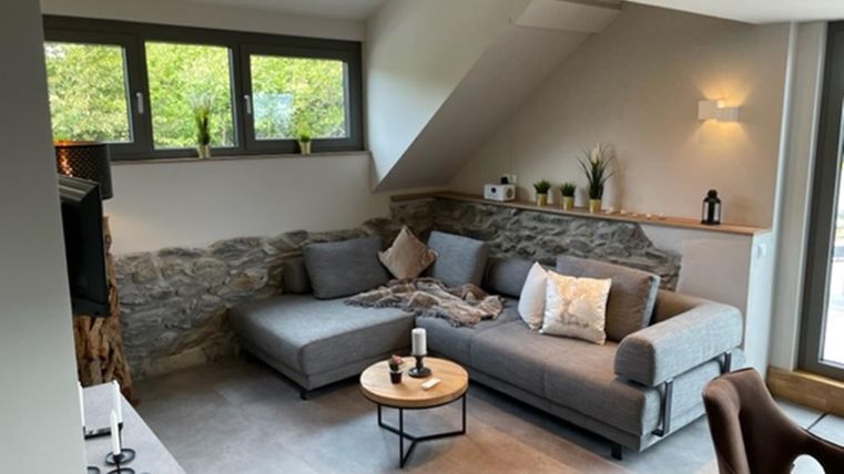 Een moderne woonkamer met een grijze bank en een ronde tafel. Grote ramen zorgen voor veel licht en een uitzicht op de groene omgeving.
