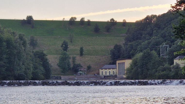 A tranquil river with a wall in the foreground. In the background, hills and buildings surrounded by trees can be seen.