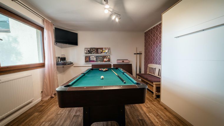 Een biljarttafel staat in een lichte kamer met een houten vloer. Op de achtergrond zijn planken en een televisie zichtbaar.