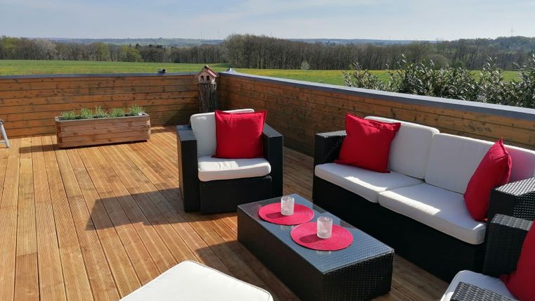 A cozy terrace with modern seating and red cushions. In the background, a green landscape stretches under a clear sky.