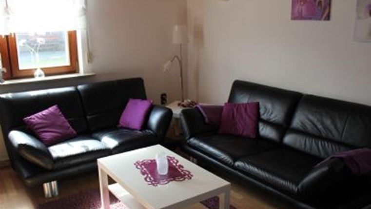 A cozy living room with two black sofas and purple cushions. A small table is in the center, on which a candle stands.