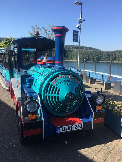 Een kleurrijk geverfde trein staat aan de oever van een water. De omgeving is groen en zonnig.