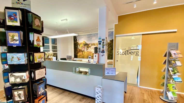 A modern reception area with a reception counter and an information stand. On the left side, postcards and brochures are displayed.