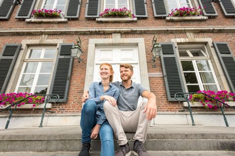 A couple is sitting on the steps in front of a beautiful building with shutters. The facade is adorned with colorful flowers.