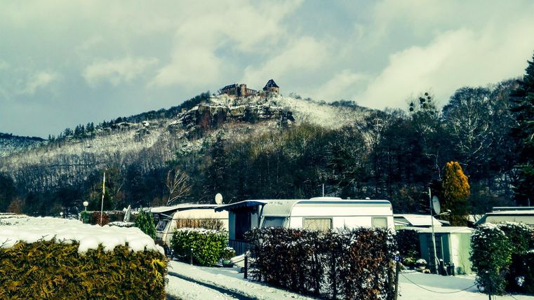 Eine verschneite Landschaft mit einem Hügel und Überresten einer Burg. Im Vordergrund sind Wohnwagen und Büsche zu sehen.