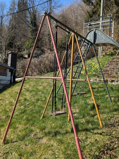 A playground with a seesaw and a slide in a green garden. In the background, there are trees and a blue sky.