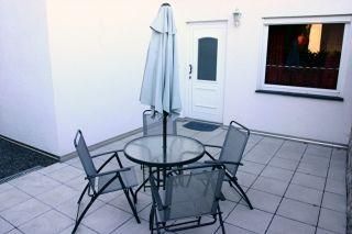 A cozy outdoor area with a table and four chairs. A sunshade stands on the terrace in front of a white wall.