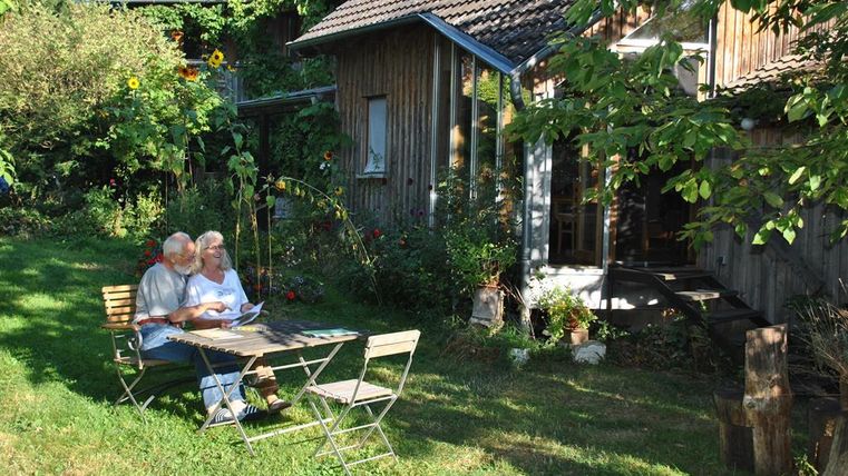 Een ouder echtpaar zit gezellig aan een tafel in de tuin en leest. Omringd door planten en een houten huis straalt de scène rust en ontspanning uit.